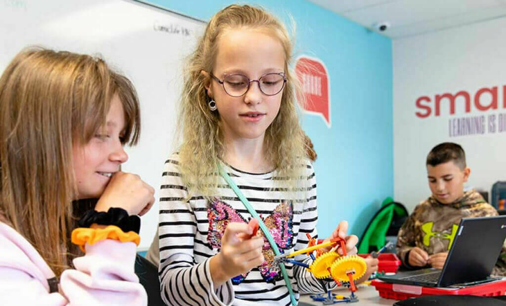 blonde girl with glasses plays with plastic gears with another blonde female classmate in a classroom setting