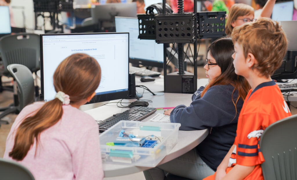 three students sit at a computer desk together viewing the screen together