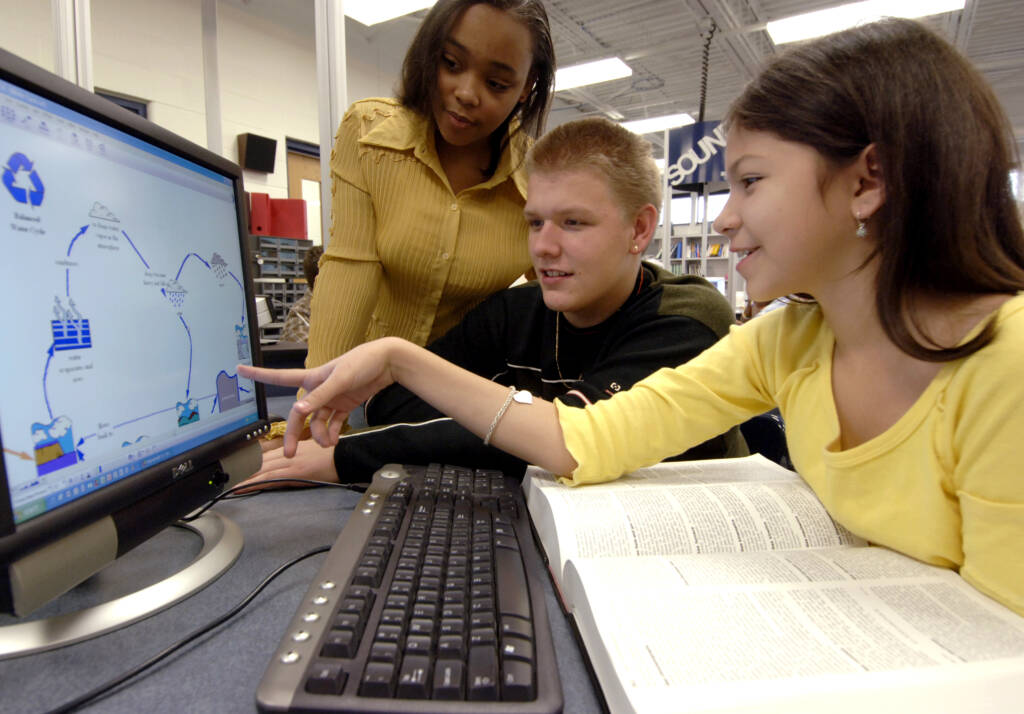 students work together on a desktop computer, pointing to the screen in collaboration